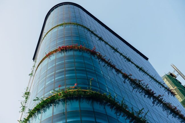 Glasfasad med växtlighet och blommande terrasser mot blå himmel